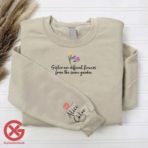 May include: A beige sweatshirt with the text "Sister are different flowers from the same garden" and a small floral design. The left sleeve has a pink heart and the text "Alice Chloe" in black.