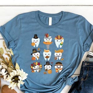 May include: A light blue t-shirt featuring a graphic of cartoon teeth dressed in Thanksgiving attire. The design includes pumpkins, hats, and fall leaves. The shirt is displayed with jeans, shoes, and decorative pumpkins.