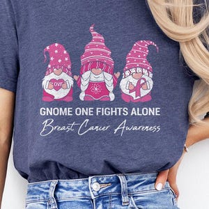 May include: Navy blue t-shirt featuring three pink gnome characters with the text "Gnome One Fights Alone Breast Cancer Awareness". The gnomes have pink hats and hold a heart, a ribbon, and a breast cancer awareness ribbon.