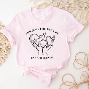 May include: Pink t-shirt with a black and white graphic of hands holding a baby with the text "Holding the future in our hands".