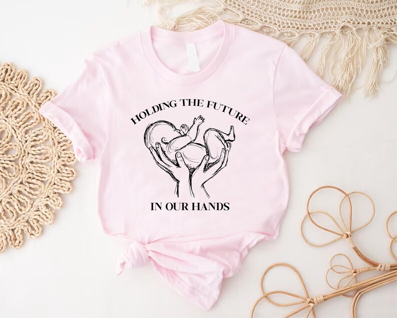 May include: Pink t-shirt with a black and white graphic of hands holding a baby with the text "Holding the future in our hands".