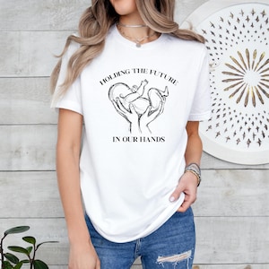 May include: White t-shirt with a black graphic of two hands holding a baby in a heart shape. The text "Holding the future in our hands" is printed below the graphic.