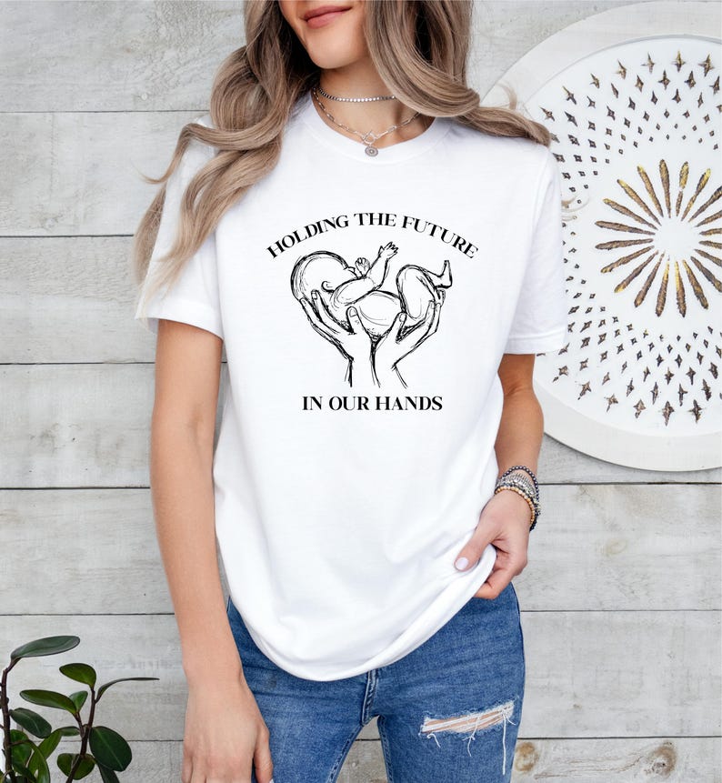 May include: White t-shirt with a black graphic of two hands holding a baby in a heart shape. The text "Holding the future in our hands" is printed below the graphic.