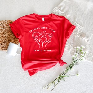 May include: Red t-shirt with a white graphic of two hands holding a baby in a heart shape. The text "Holding the future in our hands" is printed below the graphic.