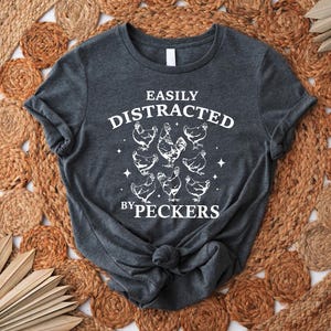 Op de afbeelding: Een donkergrijs T-shirt met de tekst "Easily Distracted by Peckers" in wit. De tekst wordt omringd door een cirkel van witte kippen.