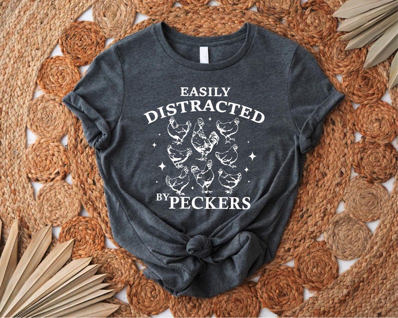 Op de afbeelding: Een donkergrijs T-shirt met de tekst "Easily Distracted by Peckers" in wit. De tekst wordt omringd door een cirkel van witte kippen.