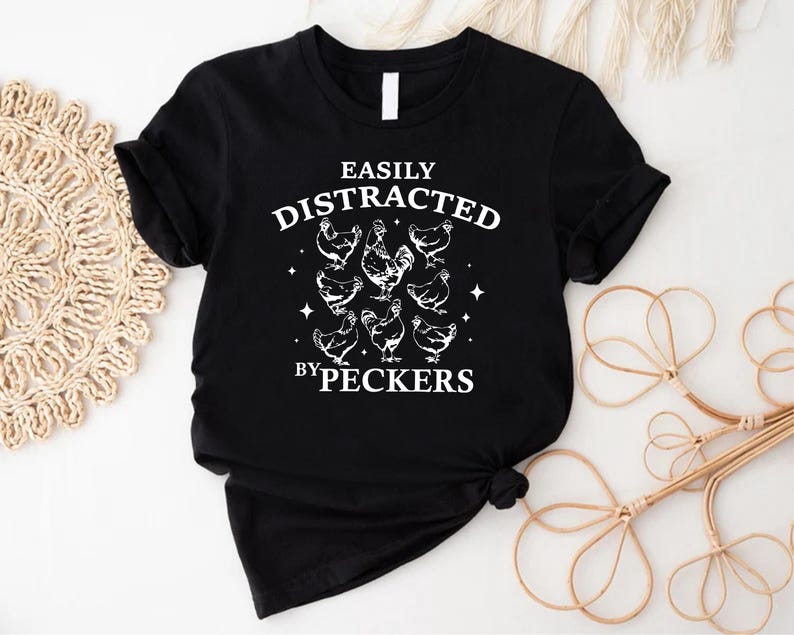 Op de afbeelding: Zwart T-shirt met witte tekst die "Easily Distracted by Peckers" luidt en een afbeelding van kippen.