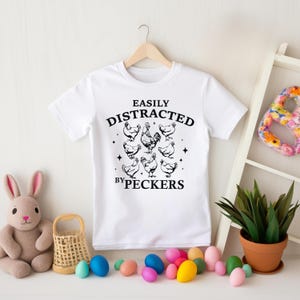 Op de afbeelding: Wit T-shirt met zwarte tekst die "Easily Distracted by Peckers" luidt en een afbeelding van kippen.