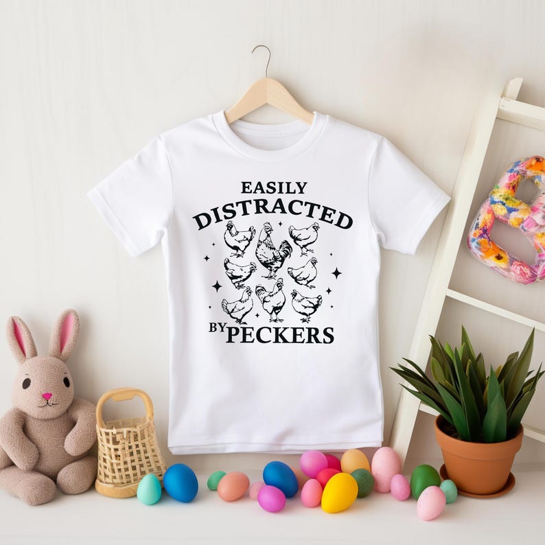 Op de afbeelding: Wit T-shirt met zwarte tekst die "Easily Distracted by Peckers" luidt en een afbeelding van kippen.