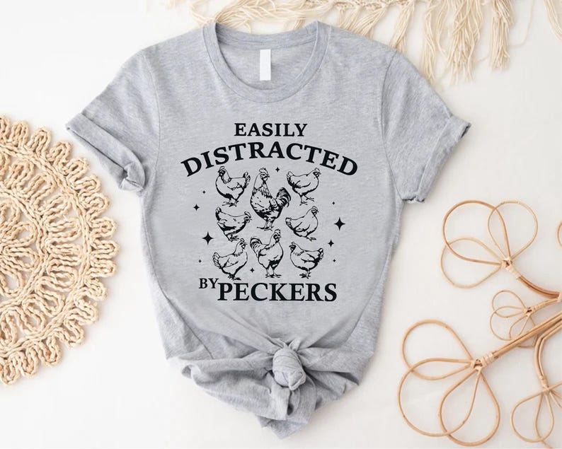 Op de afbeelding: Grijs T-shirt met een zwarte afbeelding die "Gemakkelijk afgeleid door pikken" zegt en een haan en kippen toont.