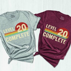 May include: Two t-shirts with a retro-style graphic that says "Level 20 Complete". One shirt is gray and the other is maroon.