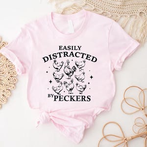 Op de afbeelding: Roze T-shirt met zwarte tekst die "Easily Distracted by Peckers" zegt en een afbeelding van kippen.