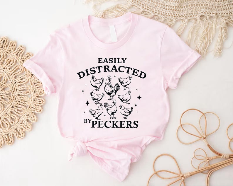 Op de afbeelding: Roze T-shirt met zwarte tekst die "Easily Distracted by Peckers" zegt en een afbeelding van kippen.