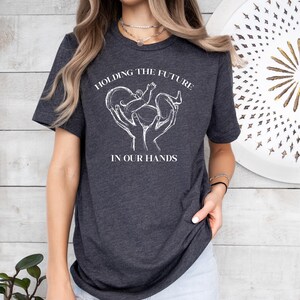 May include: A dark grey t-shirt with a white graphic of hands holding a baby with the text "Holding the Future in Our Hands".