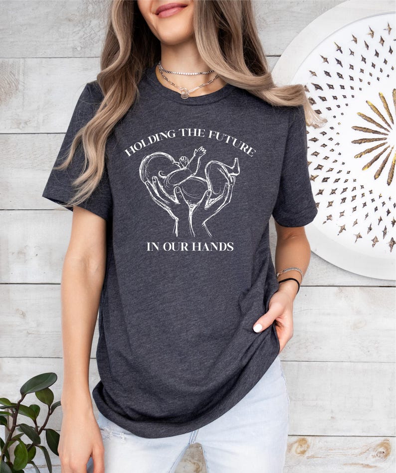 May include: A dark grey t-shirt with a white graphic of hands holding a baby with the text "Holding the Future in Our Hands".