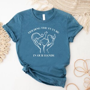 May include: A teal blue t-shirt with white text that reads "Holding the future in our hands". The text is centered on the shirt and is surrounded by a white outline of a heart with a baby inside.