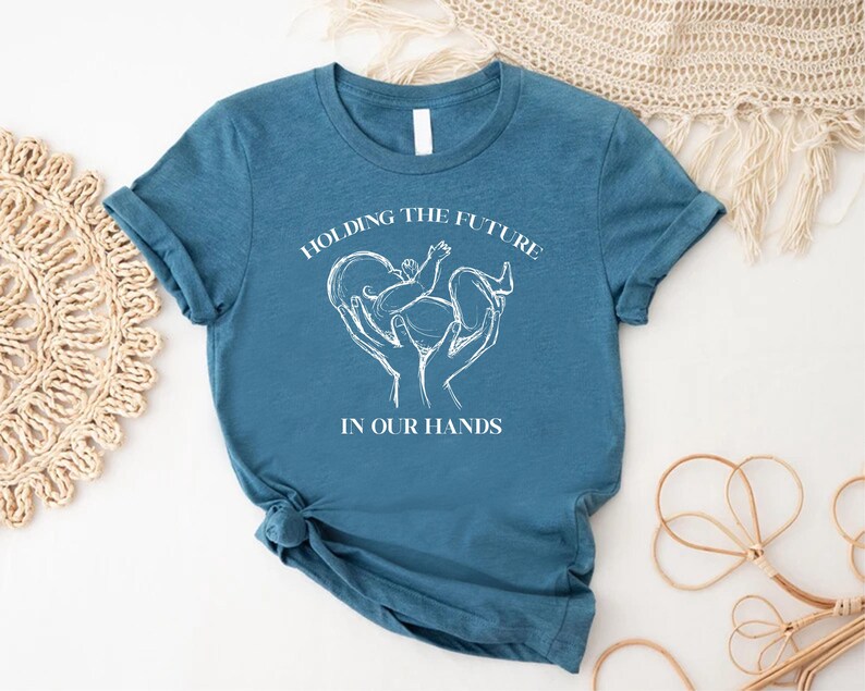 May include: A teal blue t-shirt with white text that reads "Holding the future in our hands". The text is centered on the shirt and is surrounded by a white outline of a heart with a baby inside.