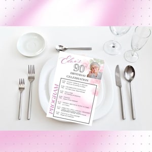 May include: A table setting with a white plate, silverware, and a printed program for Edna's 90th Birthday Celebration. The program includes a photo and schedule of events. Pink floral accents and a pink border frame the scene.