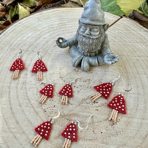 May include: Six pairs of red and white beaded mushroom earrings with beige stems hanging on a wooden surface in front of a meditating gnome figurine.