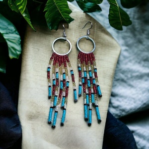 Peut inclure: Une paire de boucles d'oreilles en perles avec des cerceaux argentés. Les boucles d'oreilles présentent des brins de perles rouges, dorées, turquoise et noires. Les boucles d'oreilles sont présentées sur un tissu beige avec des feuilles vertes en arrière-plan.
