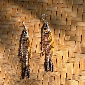 May include: A pair of long, beaded earrings with a brown, gold, and white color scheme. The earrings are made of small, round beads that are strung together in a cascading pattern.