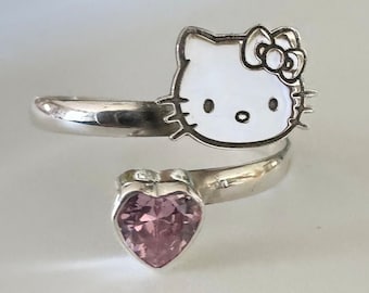 May include: A silver-toned wrap ring featuring a white enamel Hello Kitty face with a pink bow and a heart-shaped pink gemstone. The ring wraps around the finger, with the Kitty face at the top and the heart at the bottom.