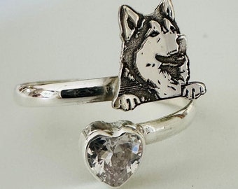 May include: A silver ring with a husky dog design. The ring wraps around the finger and features a heart-shaped clear gemstone. The husky is depicted with detailed fur. The ring is made of silver.