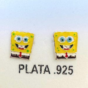 May include: A pair of silver earrings featuring a yellow cartoon character with blue eyes, a red tie, and a smiling mouth. The earrings are square-shaped and have the text "PLATA .925" below them.
