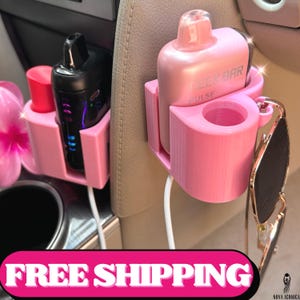 May include: Pink car accessories, including a holder for a vape device and a cup holder, are shown. The holder is designed to attach to a car's console. The image also shows a pair of sunglasses.
