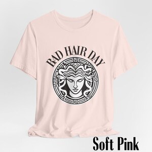 Puede incluir: Una camiseta rosa suave con un gr&aacute;fico en blanco y negro de una cabeza de Medusa dentro de un c&iacute;rculo con el texto "Bad Hair Day".