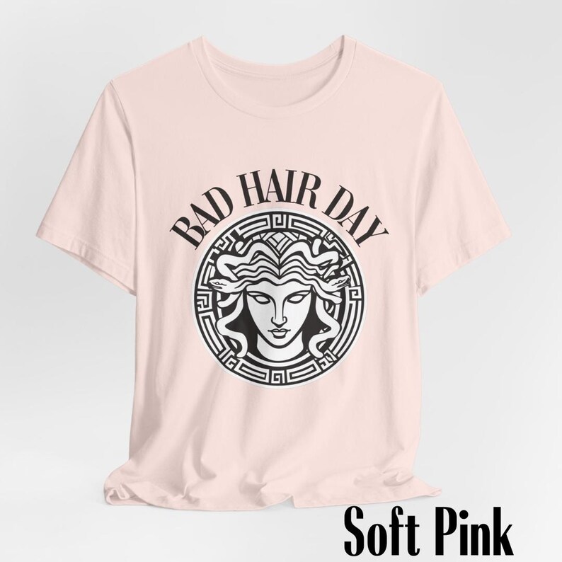 Puede incluir: Una camiseta rosa suave con un gr&aacute;fico en blanco y negro de una cabeza de Medusa dentro de un c&iacute;rculo con el texto "Bad Hair Day".