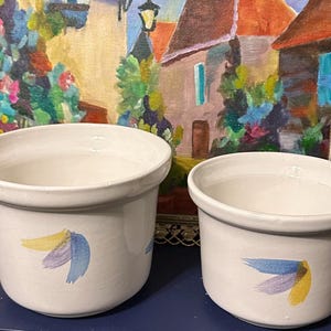 May include: Two white ceramic flower pots with blue and yellow painted designs. The pots are sitting on a dark surface in front of a framed painting.