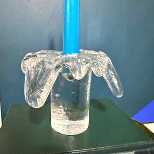 May include: A clear glass candle holder with a unique, wavy design. The holder is shaped like a flower with a cylindrical base. A blue candle is placed in the holder.