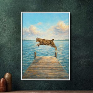 Op de afbeelding: Een ingelijste kunstdruk toont een tabby kat die van een houten pier over blauw water springt. De achtergrond heeft een blauwe lucht met wolken. Het kunstwerk is in een schilderstijl, met zichtbare penseelstreken.