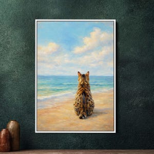 Może przedstawiać: Oprawiony obraz przedstawia kota z brązowymi i czarnymi oznaczeniami, siedzącego na piaszczystej plaży, zwróconego w stronę morza. Niebo jest niebieskie z białymi chmurami, a woda turkusowa. To spokojny krajobraz.