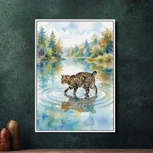 Op de afbeelding: Waterverfschilderij van een Bengaalse kat die in een meer loopt. De kat heeft een bruin-zwarte gevlekte vacht. Het water weerspiegelt de kat, de bomen en de lucht. Het kunstwerk is in een witte lijst geplaatst.