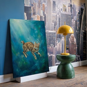 Op de afbeelding: Olieverfschilderij van een kat die onder water zwemt, met een blauwgroene achtergrond. De kat heeft bruine en zwarte vlekken. Een gele lamp en een groene kruk zijn ook zichtbaar.