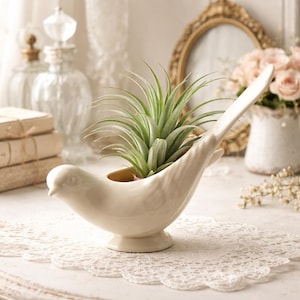 Pode incluir: Um vaso de cerâmica creme em forma de pássaro com uma planta aérea verde. O pássaro tem uma cauda longa e está pousado em um pequeno pedestal. O vaso está em uma toalha de renda branca, com outros itens decorativos ao fundo.