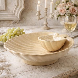 Puede incluir: Un juego de fuentes de cerámica beige, que incluye un cuenco grande con forma de concha y un cuenco más pequeño para salsas a juego. Los cuencos tienen un diseño texturizado y acanalado. El juego está sobre una mesa de madera rústica blanqueada, lo que sugiere un tema costero o de granja.