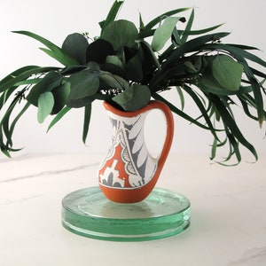 Puede incluir: Una jarra de terracota con un diseño geométrico blanco y gris, llena de hojas de eucalipto verdes, se asienta sobre un pedestal de vidrio transparente.