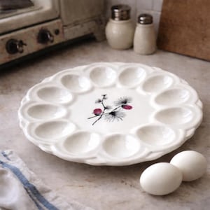 Peut inclure: Un plat de service à œufs en céramique blanche avec des bords festonnés et un motif floral au centre. Le plat comporte 12 cavités en forme d'œuf. Deux œufs blancs sont à côté du plat.