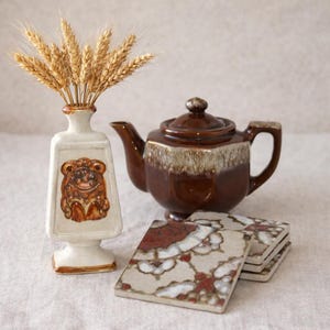 Puede incluir: Naturaleza muerta con una tetera marrón, un jarrón blanco con un relieve marrón y una pila de posavasos cuadrados con un estampado floral. El jarrón contiene espigas de trigo. El conjunto tiene una estética vintage.