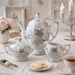 May include: A white porcelain tea set with silver accents, including a teapot, sugar bowl, and creamer. The set is decorated with a silver floral pattern and the words "25th Anniversary". A plate of cookies and a cup of tea complete the scene.
