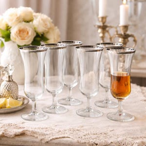 May include: A set of clear stemmed glasses with silver rims, one filled with amber liquid. The glasses are arranged on a lace tablecloth, with a vase of white roses in the background. A decorative perfume bottle and lemon slices are also present.