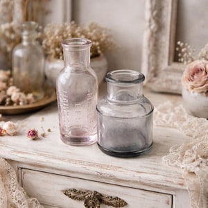 May include: Two antique glass bottles, one pink and one gray, sit on a white surface. The pink bottle has text on it. Dried flowers and lace add to the vintage aesthetic. The bottles are likely for decorative purposes.