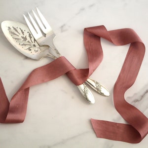 May include: A pair of silver serving utensils tied together with a dusty rose satin ribbon.