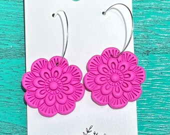 May include: A pair of bright pink flower-shaped earrings with silver hoops. The earrings are made of clay and have detailed floral patterns. The card behind the earrings reads "CLAY BY KIRSTIN HANDMADE IN LEESBURG, FL."