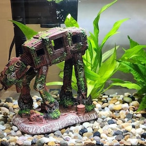 K&ouml;nnte beinhalten: Detaillierte Aquariumdekoration in Form eines AT-AT-Walkers aus Star Wars. Die braune und gr&uuml;ne Kunstharzfigur ist mit moosartigen Details bedeckt und steht auf einem felsigen Sockel. Gr&uuml;ne Pflanzen und Kies umgeben den Walker im Aquarium.