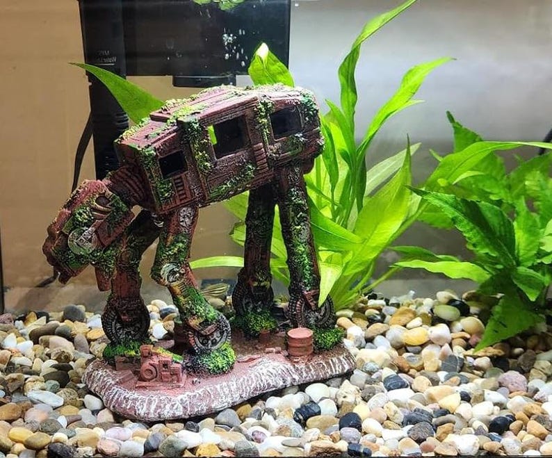 K&ouml;nnte beinhalten: Detaillierte Aquariumdekoration in Form eines AT-AT-Walkers aus Star Wars. Die braune und gr&uuml;ne Kunstharzfigur ist mit moosartigen Details bedeckt und steht auf einem felsigen Sockel. Gr&uuml;ne Pflanzen und Kies umgeben den Walker im Aquarium.