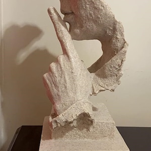 Pode incluir: Uma escultura abstrata branca de uma pessoa com o dedo nos lábios, como para dizer 'shhh'. A escultura é feita de um material áspero e texturizado e está montada em uma base quadrada.